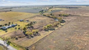 Aerial view of a rural landscape with open fields, scattered trees, a few houses, a pond, and a winding road, under a partly cloudy sky.