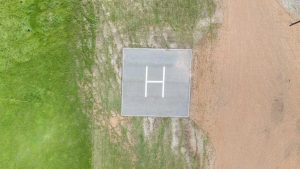 Aerial view of a concrete helipad marked with a large white "H," surrounded by grass on the left and bare dirt on the right.