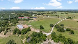 Aerial view of a rural landscape with green fields, scattered trees, a winding road, a large barn-like building, and several houses in the distance under a partly cloudy sky.