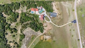 Aerial view of a large house with a pool, solar panels, and long driveway, surrounded by trees and open grassy land with a nearby road.