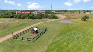 Aerial view of a fenced enclosure with a small shed on a grassy field, a dirt road leading to several buildings in the distance, trees, a pond, and power lines under a blue sky with clouds.