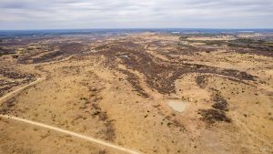 Aerial view of a vast, dry landscape with patches of sparse vegetation, small ponds, and rolling hills under a cloudy sky; a dirt road runs across the lower left corner of the image.