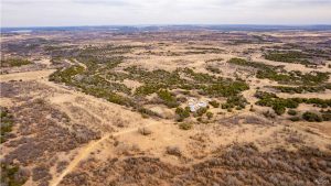 Aerial view of a sprawling, dry landscape with patches of green trees, rolling hills, and a small cluster of buildings near a pond in the center. Dirt roads and natural brush are scattered throughout the scene.