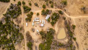 Aerial view of a rural campsite with several trailers and structures surrounded by trees, dirt roads, and a small pond on dry, brown grassland.
