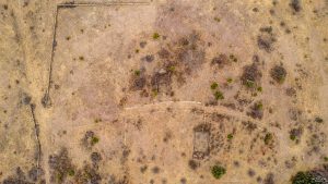 Aerial view of a dry, sparsely vegetated landscape with patches of grass and bushes, remnants of old rectangular foundations, and a faint curved dirt path crossing through the area.