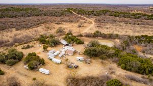 Aerial view of a rural campsite with several RVs, trailers, and small buildings arranged in a clearing surrounded by dry brush, trees, and dirt roads, with a small pond nearby. Expansive landscape visible in the background.