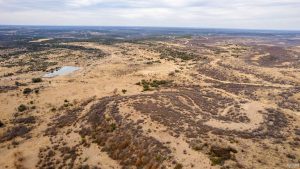Aerial view of a vast, dry landscape with patches of shrubs and grass, rolling hills, and a small pond. The sky is partly cloudy, and the land stretches into the distance.