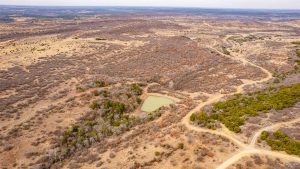 Aerial view of a dry, rugged landscape with sparse vegetation, winding dirt roads, and a small, greenish pond surrounded by trees in the center. The horizon shows rolling hills under a cloudy sky.