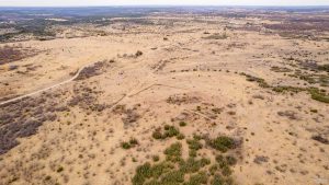 Aerial view of a vast, dry, and sparsely vegetated landscape with rolling hills, scattered bushes, and a dirt road winding through the scene under an overcast sky.