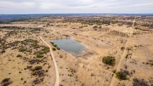 Aerial view of a dry, hilly landscape with sparse vegetation and dirt roads surrounding a small, irregularly shaped pond under a cloudy sky.