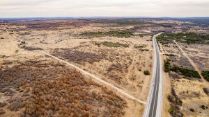 Aerial view of a rural landscape with a long, straight highway running through dry, grassy fields and patches of sparse vegetation under a cloudy sky.