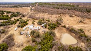 Aerial view of a rural campsite with RVs, small buildings, scattered trees, and a pond, surrounded by dry fields and rolling hills under an overcast sky.
