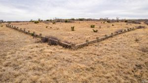 A V-shaped stone wall encloses a dry, grassy field with sparse shrubs under a cloudy sky. The wall is built from stacked rocks and curves gently at the corner.