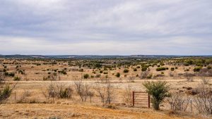 A wide, open landscape with dry grass, scattered bushes, and a few bare trees under a cloudy sky. A rusty metal gate stands in the foreground, leading toward rolling hills in the distance.