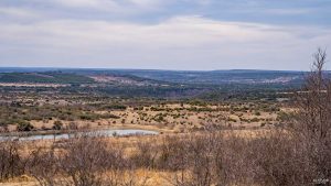 A wide, dry landscape with sparse shrubs and trees, a small pond in the foreground, and rolling hills under a cloudy sky in the distance.