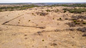 Aerial view of a dry, grassy landscape with sparse shrubs and bushes, bordered by a low, weathered fence that zigzags across the terrain. Rolling hills and patches of greenery are visible in the distance under an overcast sky.
