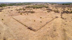Aerial view of a dry, grassy landscape with a V-shaped stone wall enclosure in the center, surrounded by sparse vegetation and rolling hills in the background.