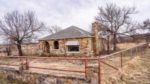 An old stone house with a shingled roof and arched porch sits behind a rusted wire fence in a dry, grassy field, surrounded by leafless trees under a cloudy sky.