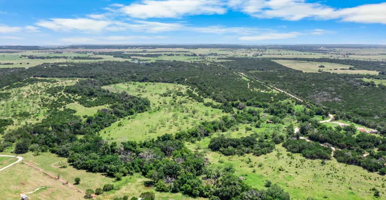 Aerial view of a vast Erath County, Texas rural landscape with green fields, scattered trees, and patches of forest under a blue sky with scattered clouds. Dirt roads and open areas are visible throughout the countryside.